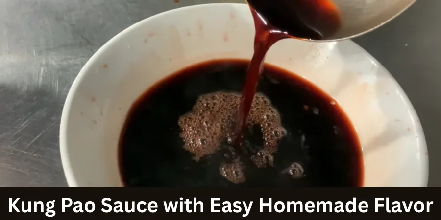 Overhead view of thick, glossy dark Kung Pao sauce in a white bowl with a wooden spoon, garnished with visible chili flakes and surrounded by dried red chilies on a light surface.